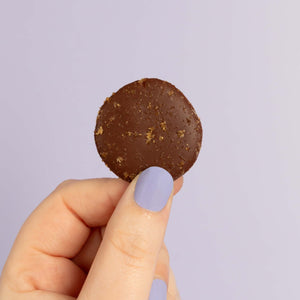 Close up of a salted caramel chocolate button, held by a finger and thumb in front of a purple backdrop.